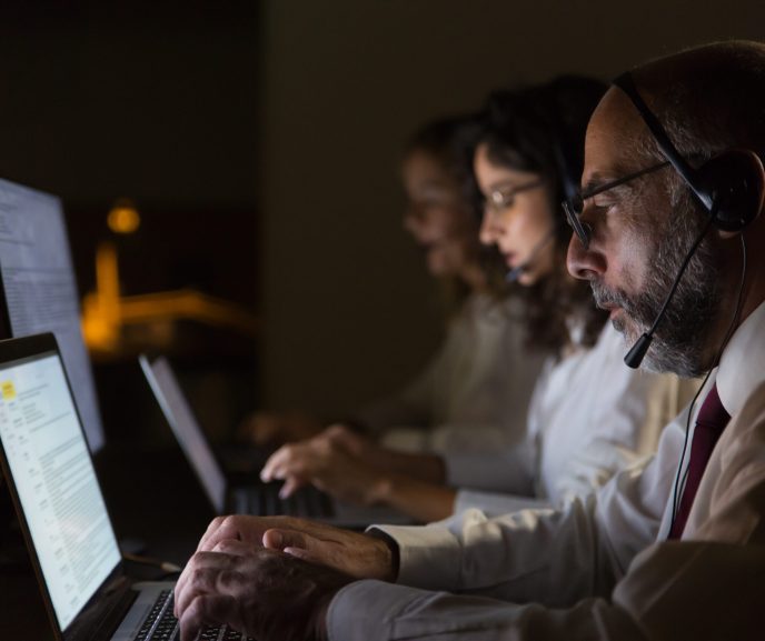 Focused coworkers in headsets typing on laptops. Side view of focused call center operators in headsets using laptop computers in dark office. Working late concept